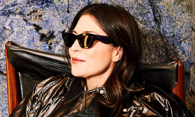 Model with dark brown hair in a leather jacket wearing nk x illesteva Donna Sunglasses Black/Cobalt with Grey