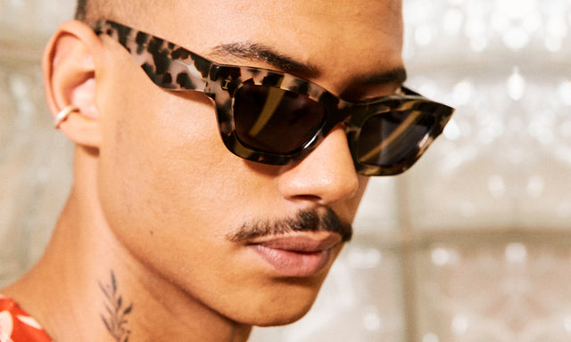 Model with a mustache and neck tattoo wearing nk x illesteva Donna Sunglasses White Tortoise with Grey