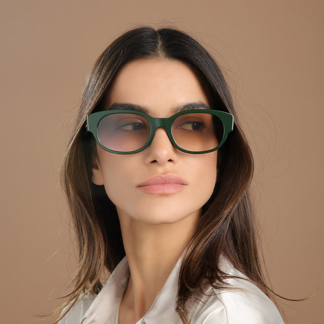 Brunette model in a cream silk blouse looking over her left shoulder wearing Montreal E Sunglasses Evergreen with Taupe Flat Gradient