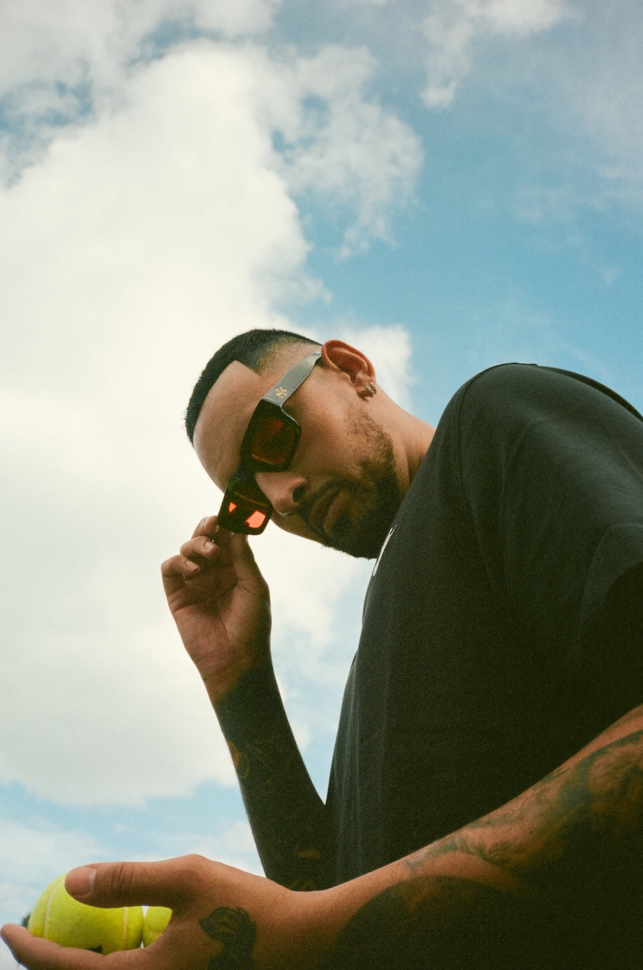 Nick Kyrgios holding a tennis ball outdoors wearing Nick Kyrgios x illesteva Sunglasses in Black with Red