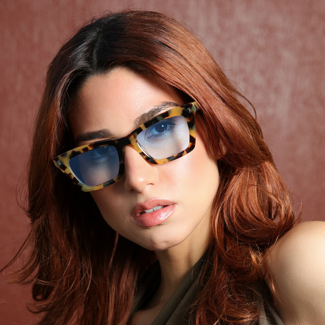 Woman near a maroon wall wearing Portugal Sunglasses Tortoise with Blue Flat Gradient See Through
