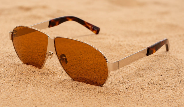 Niki Sunglasses in Silver with Desert lenses, showcased over a sandy beach