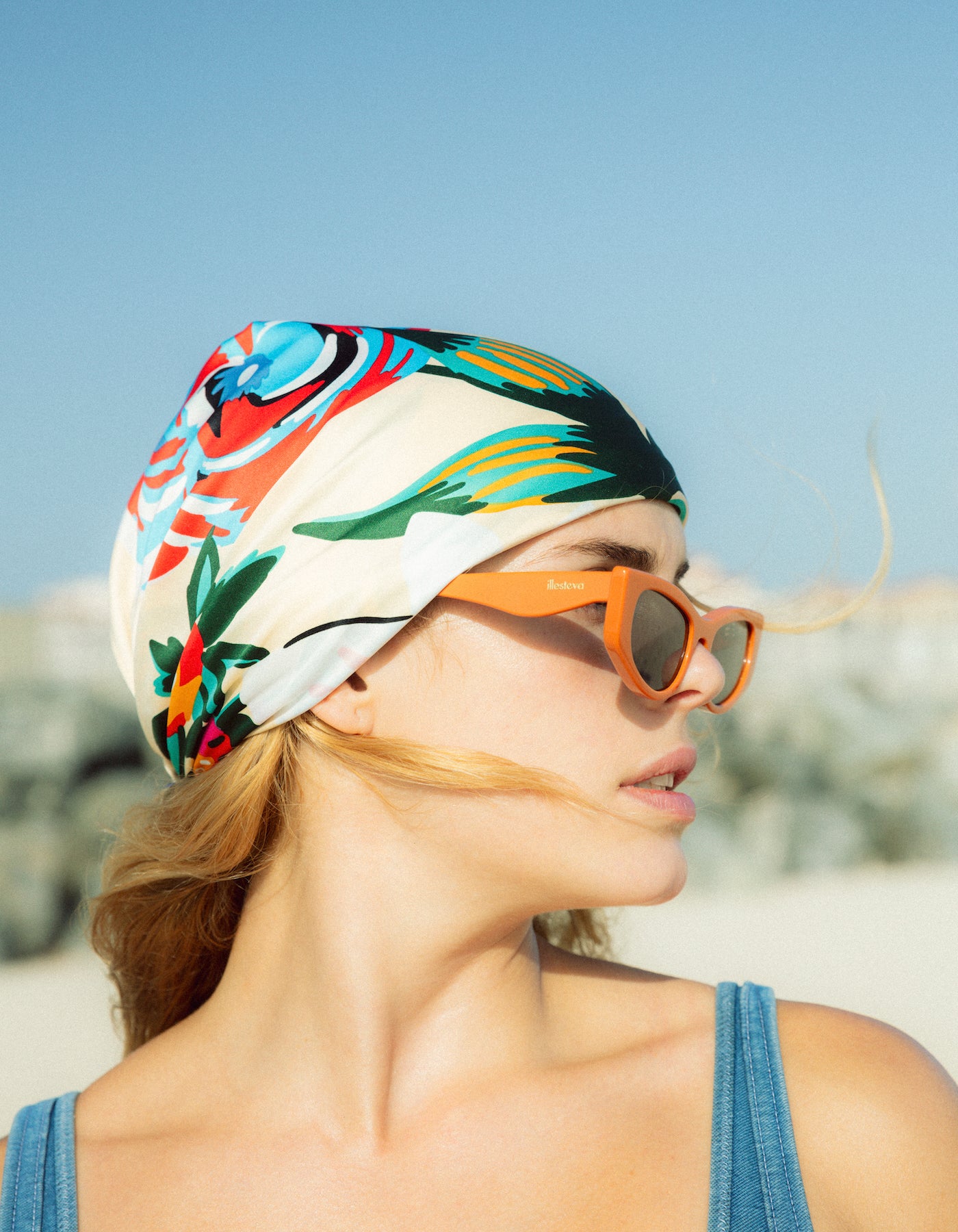 Woman wearing a colorful headscarf and Alexa Sunglasses in Aperol on a beach.