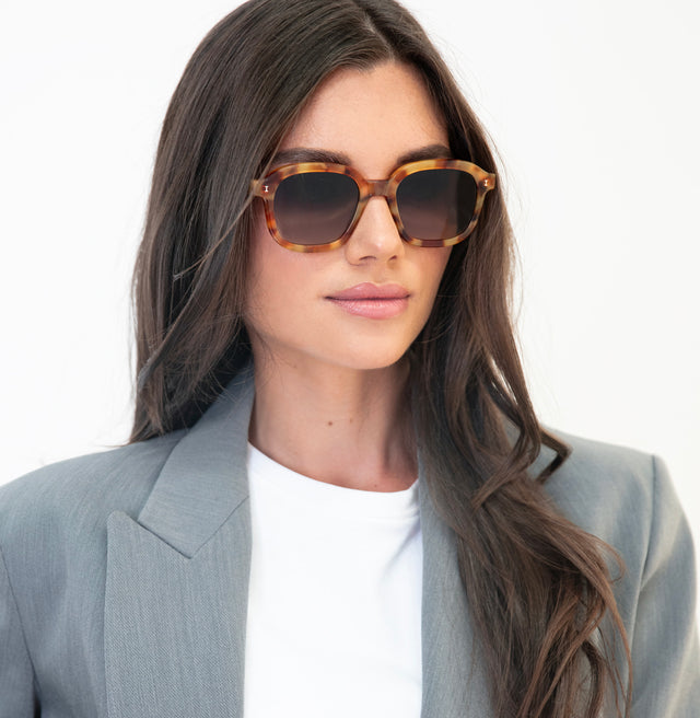 Brunette model with wavy hair in a grey blazer wearing Bogota Sunglasses Amber with Brown Flat Gradient