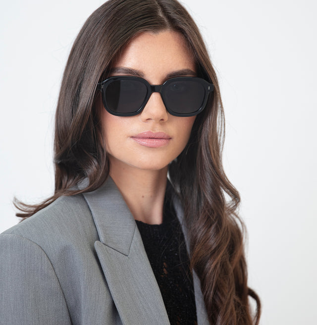 Brunette model with wavy hair in a grey blazer wearing Bogota Sunglasses Black with Grey Flat