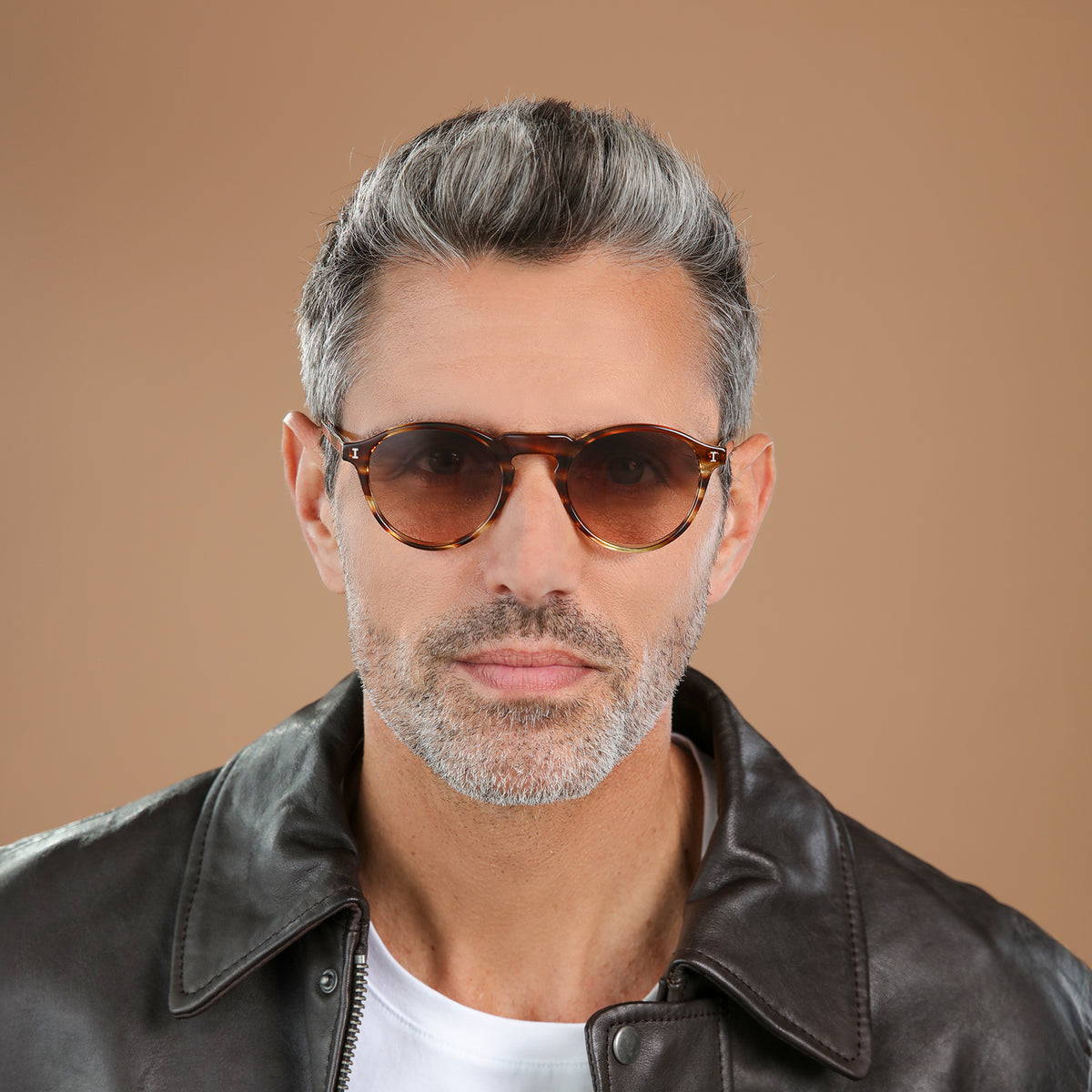 Man wearing Capri Sunglasses in Sand Dune with Brown Gradient lenses, front view, Model with salt and pepper hair and beard in a black leather jacket