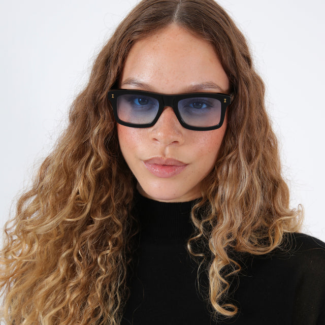 Brunette model with wavy hair in a black mockneck wearing Catania Sunglasses Black/Gold with Blue Flat Gradient See Through