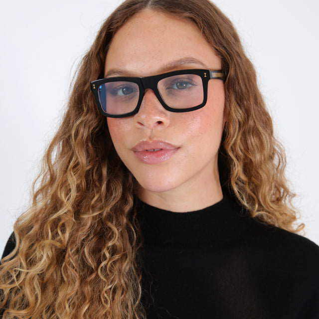 Brunette model with wavy ombre hair in a black mockneck wearing Catania Optical Black/Gold Optical