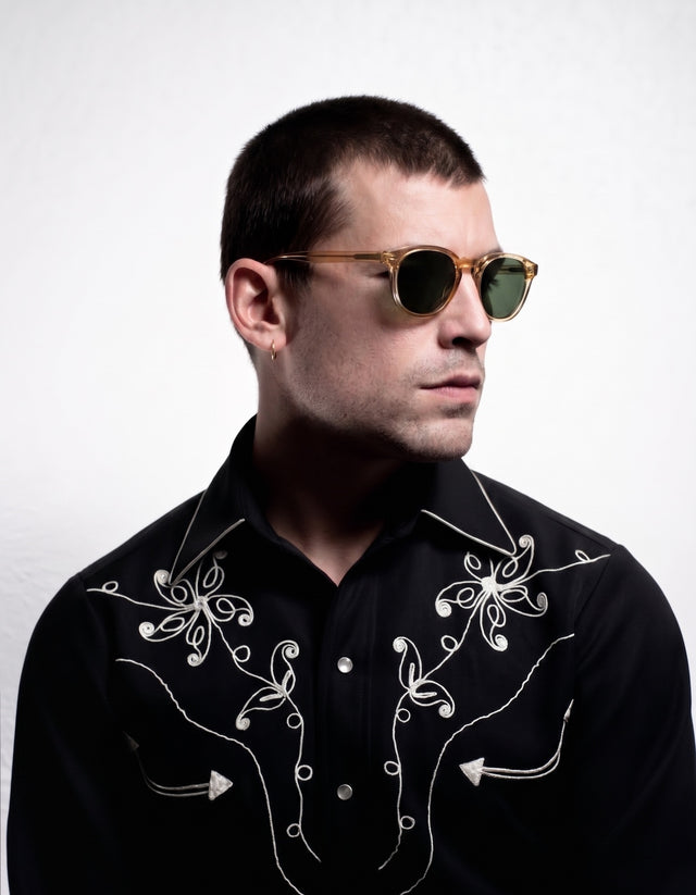 A man wearing Eldridge sunglasses in Citrine with olive lenses and a black western-style embroidered shirt, photographed against a light gray studio background.