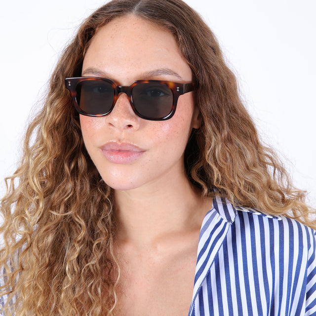 Brunette model with ombre, wavy hair in a striped blouse wearing Ellison Sunglasses Havana with Olive Flat