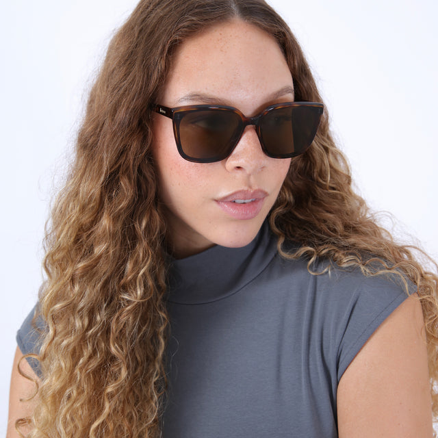Brunette model with ombre wavy hair in a gray, sleeveless turtleneck wearing Mallorca Sunglasses Havana with Brown Flat
