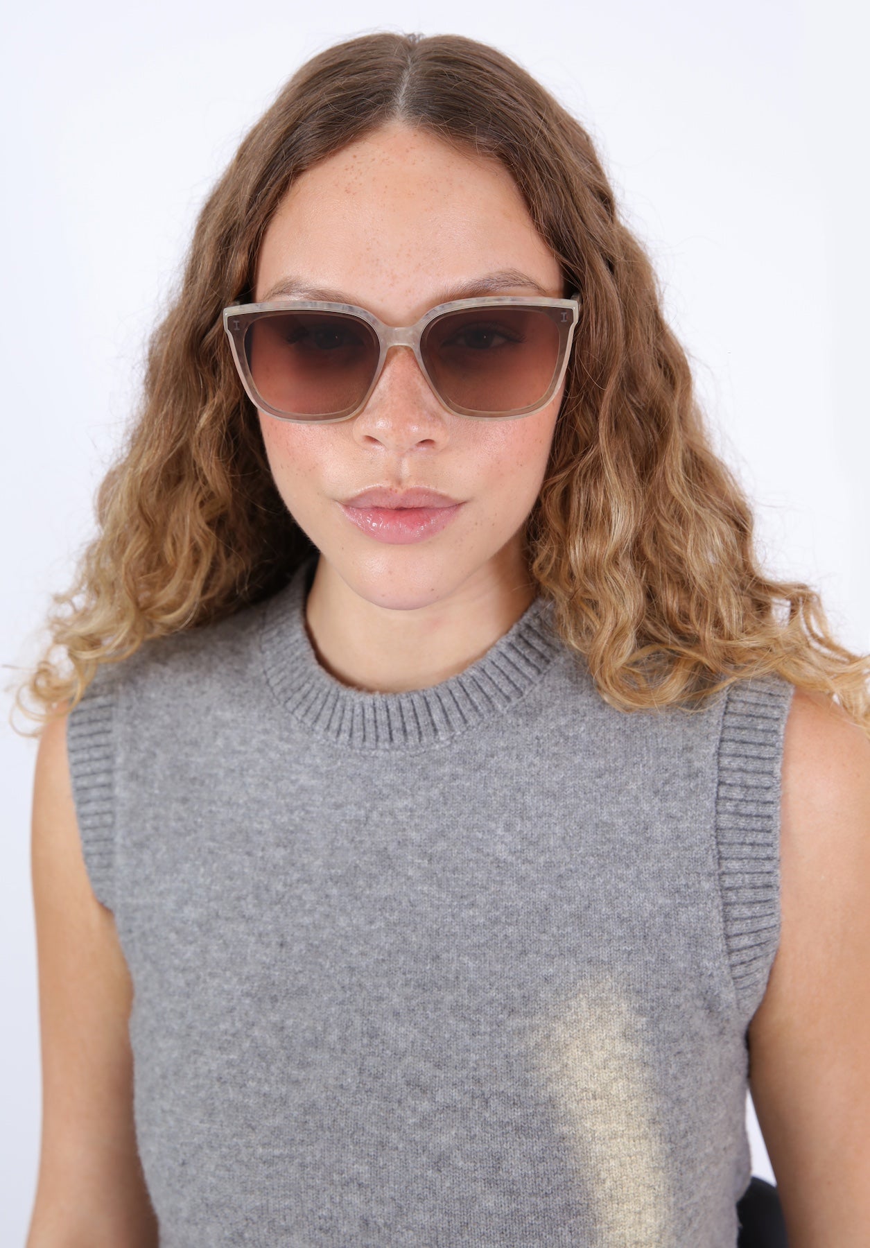 Brunette model with ombré curly hair wearing Mallorca Sunglasses in Bone paired with a grey sweater vest.