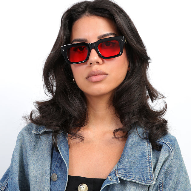 Brunette model with loose curls in a jean jacket wearing Nick Kyrgios x illesteva 2 Sunglasses Black with Red Flat See Through