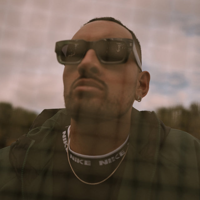 Nick Kyrgios close up through a mesh fence at a tennis court wearing Nick Kyrgios x illesteva 2 Sunglasses Pine with Grey Flat