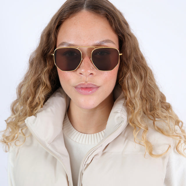 Brunette model with wavy, ombré hair with a puffer jacket wearing Patmos 58 Sunglasses Gold with Brown Flat