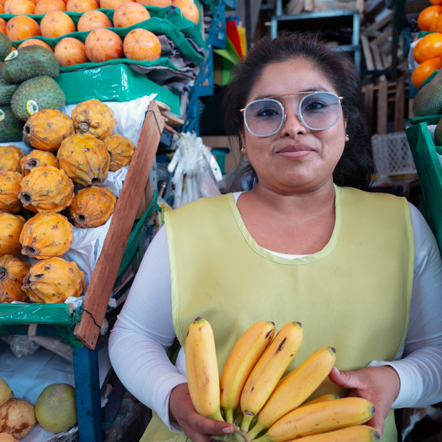 Young Peruvian woman at a market surrounded by fruit holding bananas wearing Mykonos Ace Sunglasses in Celeste