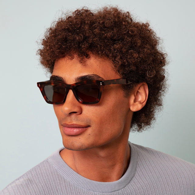 Model with afro-curly hair in a baby blue knit sweatshirt wearing Portugal Sunglasses Havana with Grey Flat