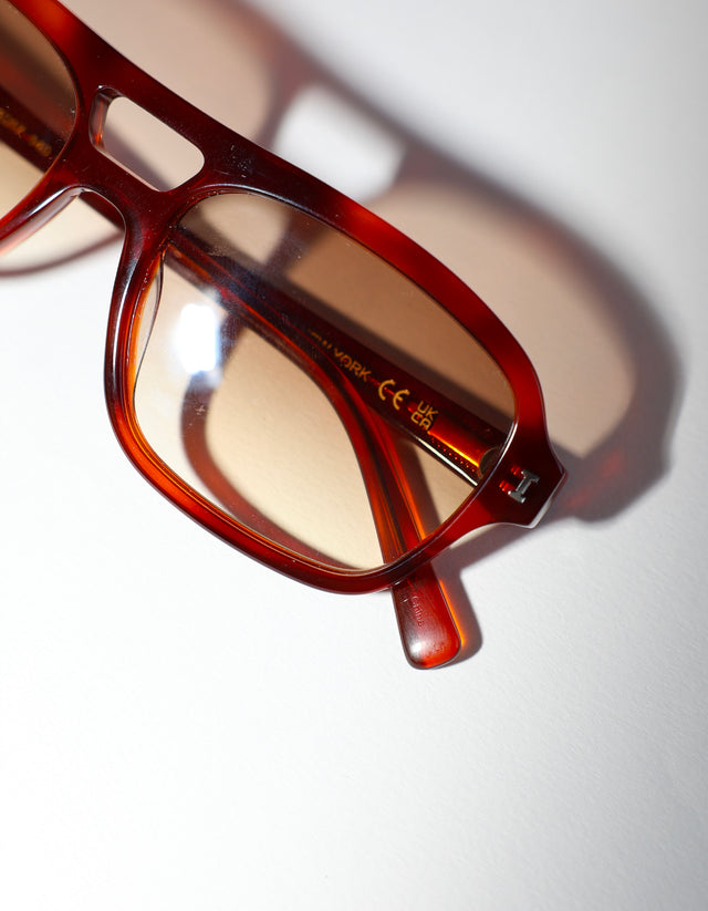 Close-up of a pair of Queens sunglasses with a translucent reddish-brown acetate frame resting on a white surface with a soft shadow.