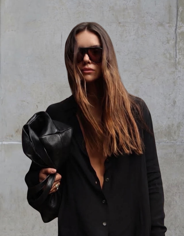 A woman with long brown hair wearing Telluride sunglasses, a black button-up shirt, and holding a black leather clutch bag near a concrete wall