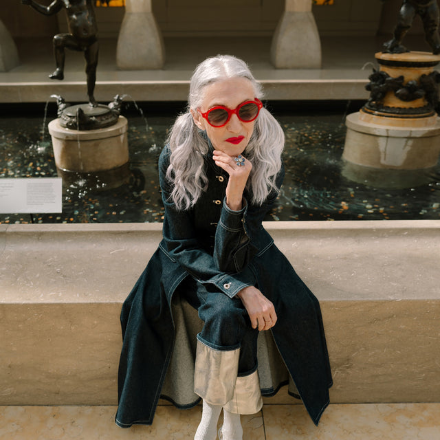 Model with silver hair sitting in front of statues wearing The Met x illesteva Sunglasses Red with Grey Gradient