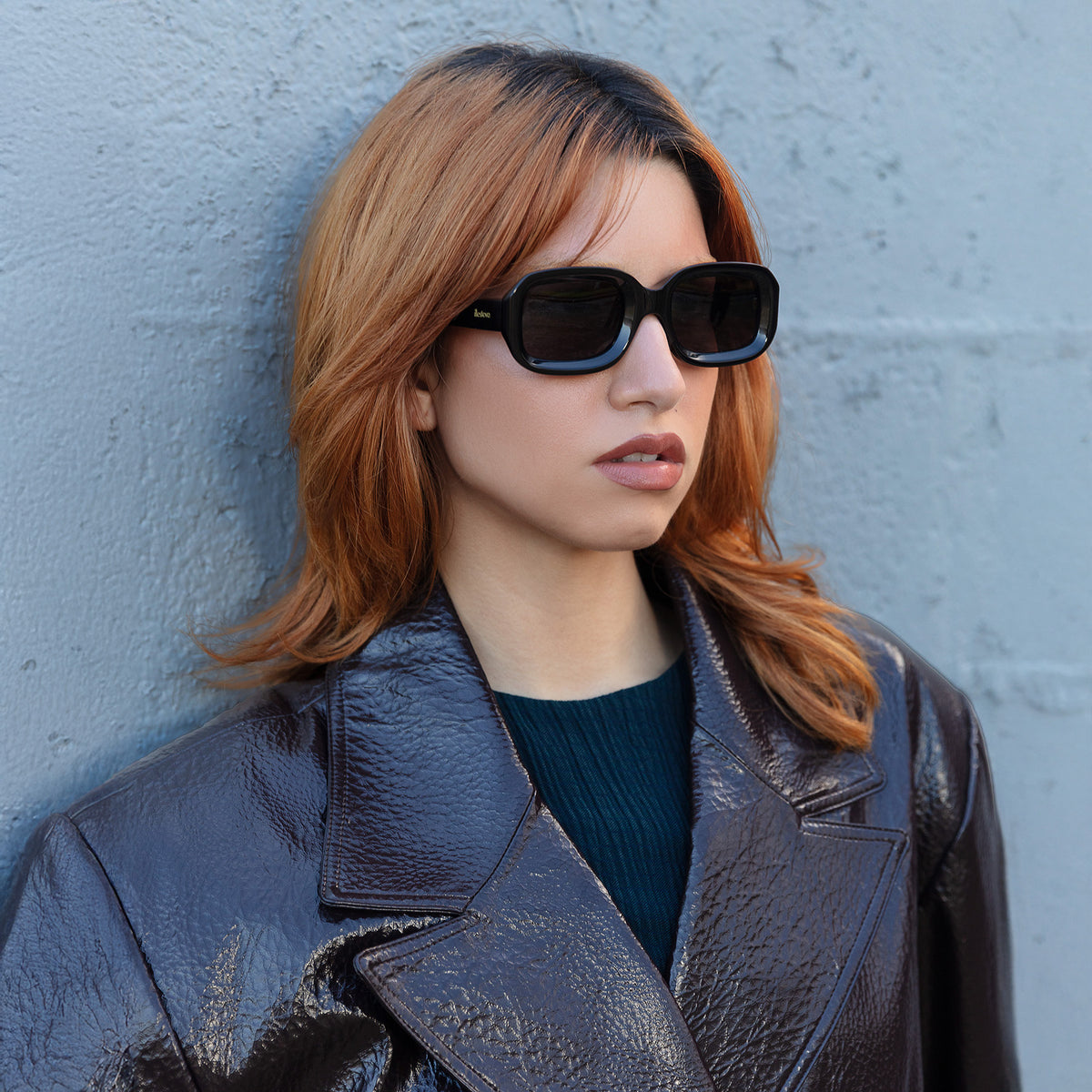 Woman wearing Vinyl Sunglasses in Black with Grey lenses, Model in a leather jacket leaning on a brick wall
